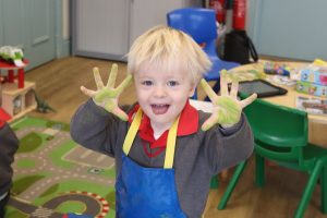 Ilkley Nursery boy smiling holding up his hands covered in green paint.