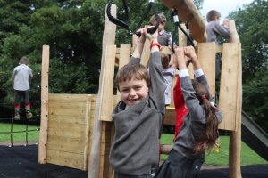 Ghyll Royd School pupil enjoys playing on new playground equipment. Boy smiling hanging onto post.